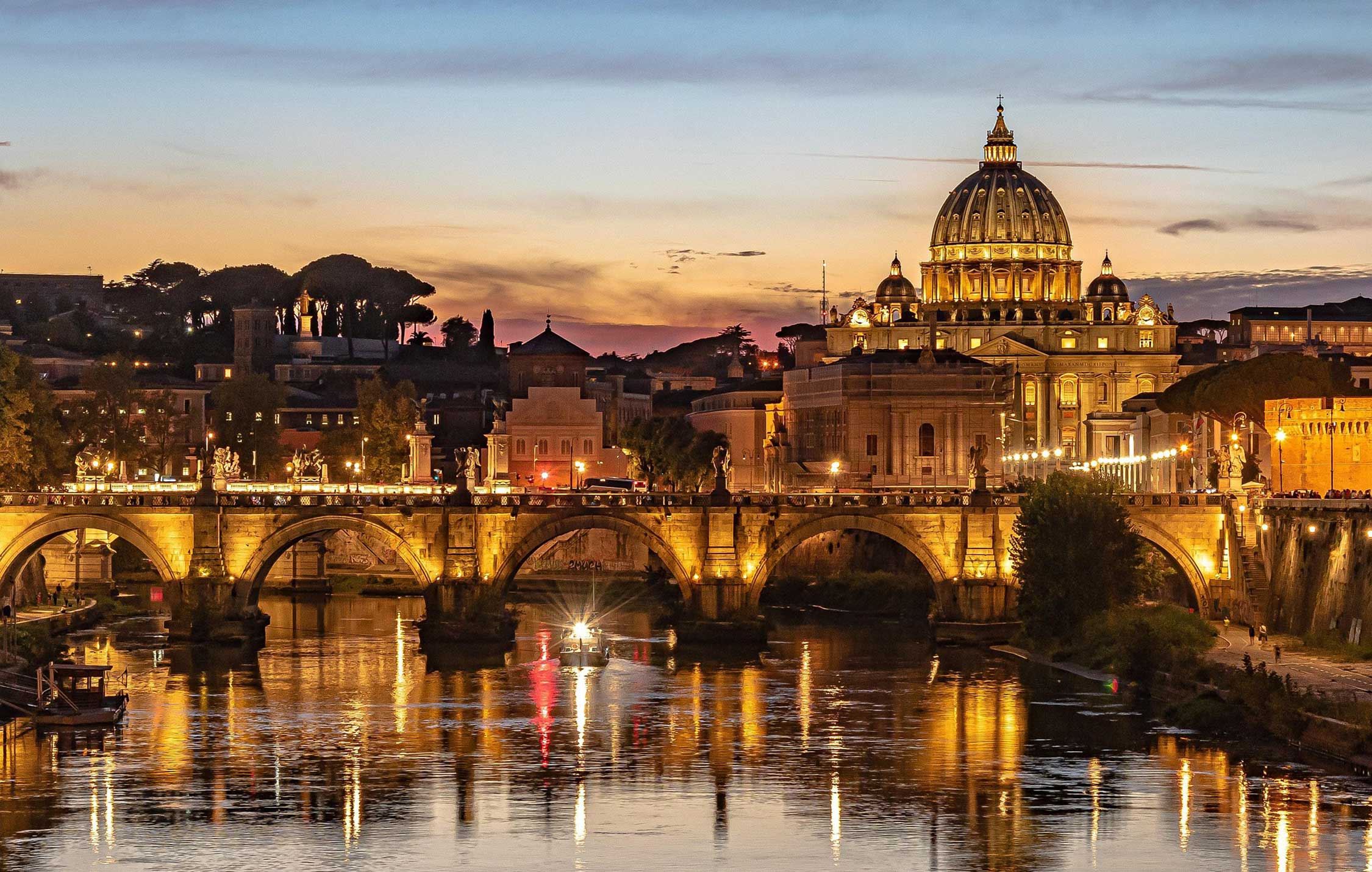 Veduta di Roma di sera, la città illuminata con i monumenti storici in controluce e il cielo scuro.